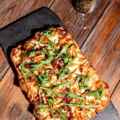 Gourmet flatbread pizza topped with arugula and melted cheese served on slate board with glass of red wine