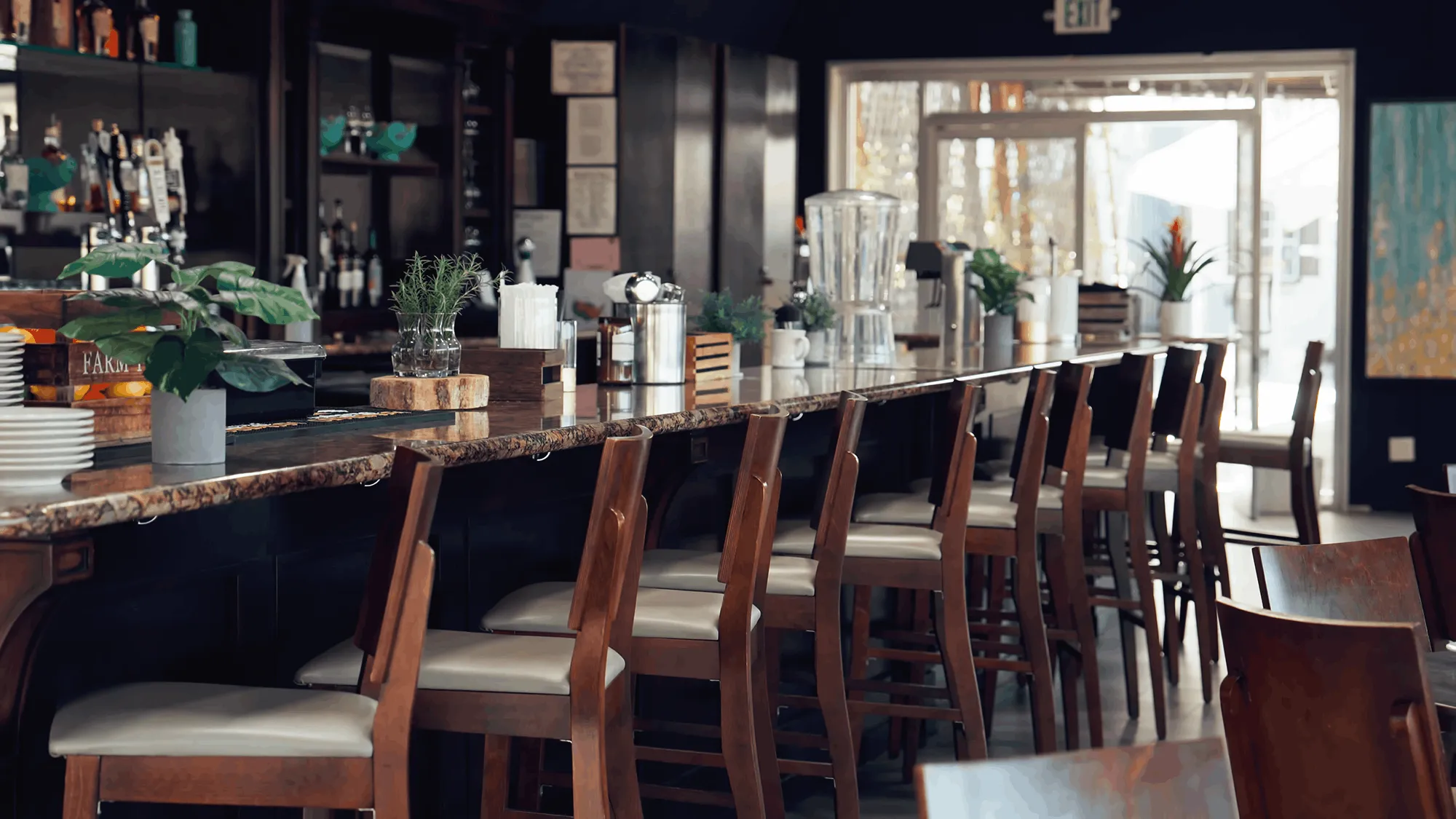 Modern restaurant bar interior with wooden high chairs, marble counter, indoor plants, and beverage shelves in the background.