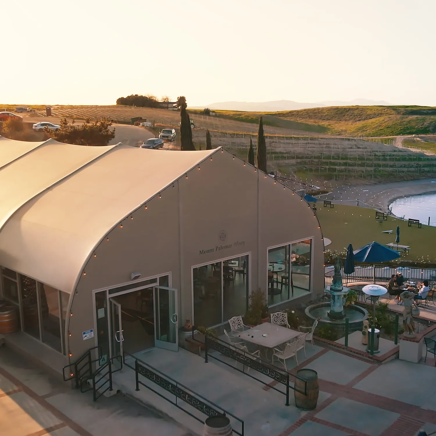 Aerial view of Mount Palomar Winery bistro building with outdoor patio seating overlooking vineyard hills and a lake at sunset