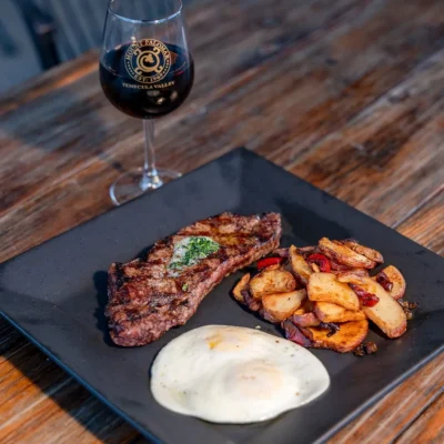 Grilled steak with sunny side egg and roasted potatoes served on black plate with red wine on wooden table